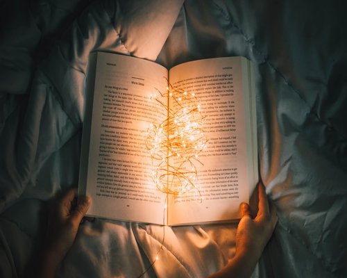 A person reading a physical book under the warm glow of a bedside lamp